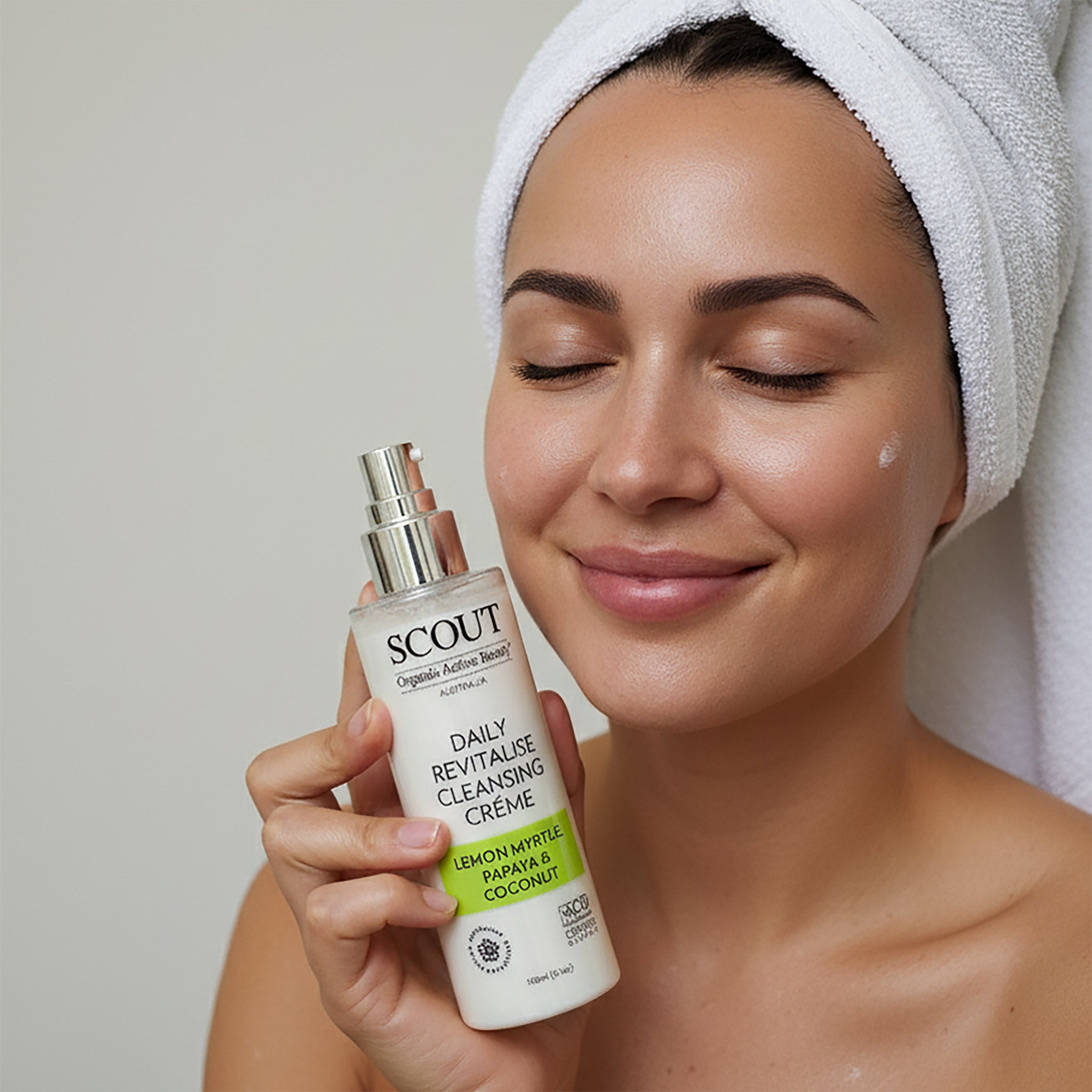 Woman holding a bottle of Scout Daily Revitalise Cleansing Creme with a towel on her head.
