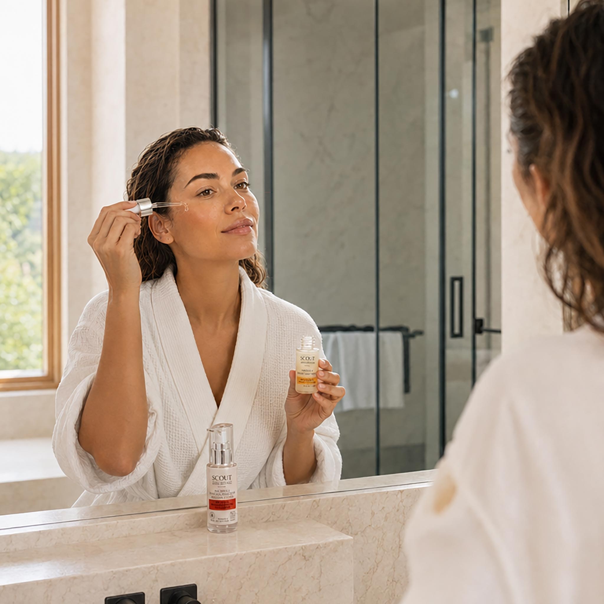 A woman in a white robe applies skincare serum from a dropper to her face while looking at her reflection in a bathroom mirror. A bottle of the same skincare product sits on the counter in front of her.