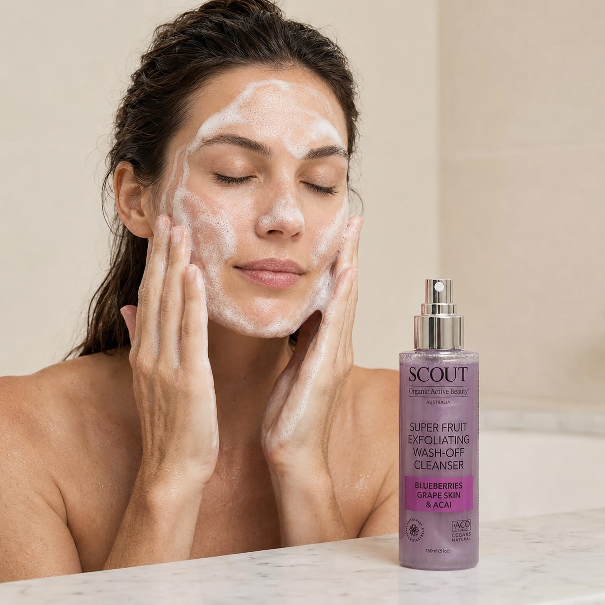 A woman with white cleanser foam on her face applies it to her cheeks while standing in a bathroom next to a bottle of SCOUT Super Fruit Exfoliating Wash-Off Cleanser.