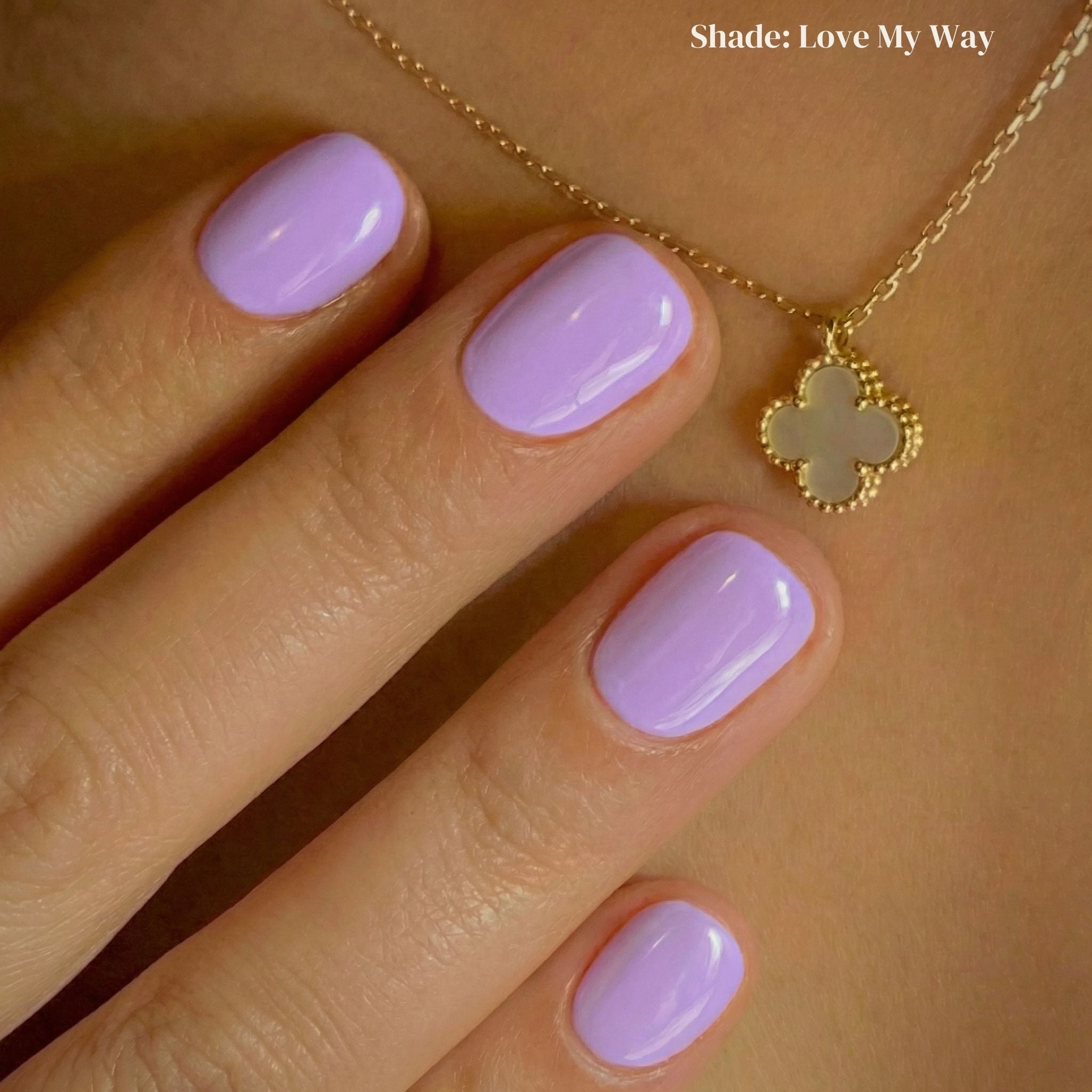 Hand with light purple nail polish wearing a gold necklace with a clover pendant on a beige background.