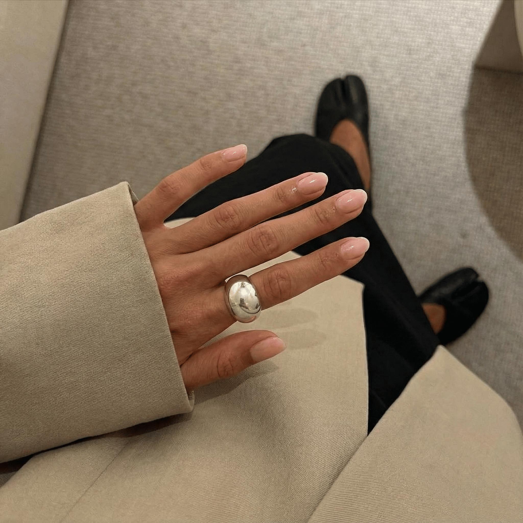 A close-up of a person's hand wearing a chunky silver ring with a polished oval stone, styled with a beige blazer and black pants.