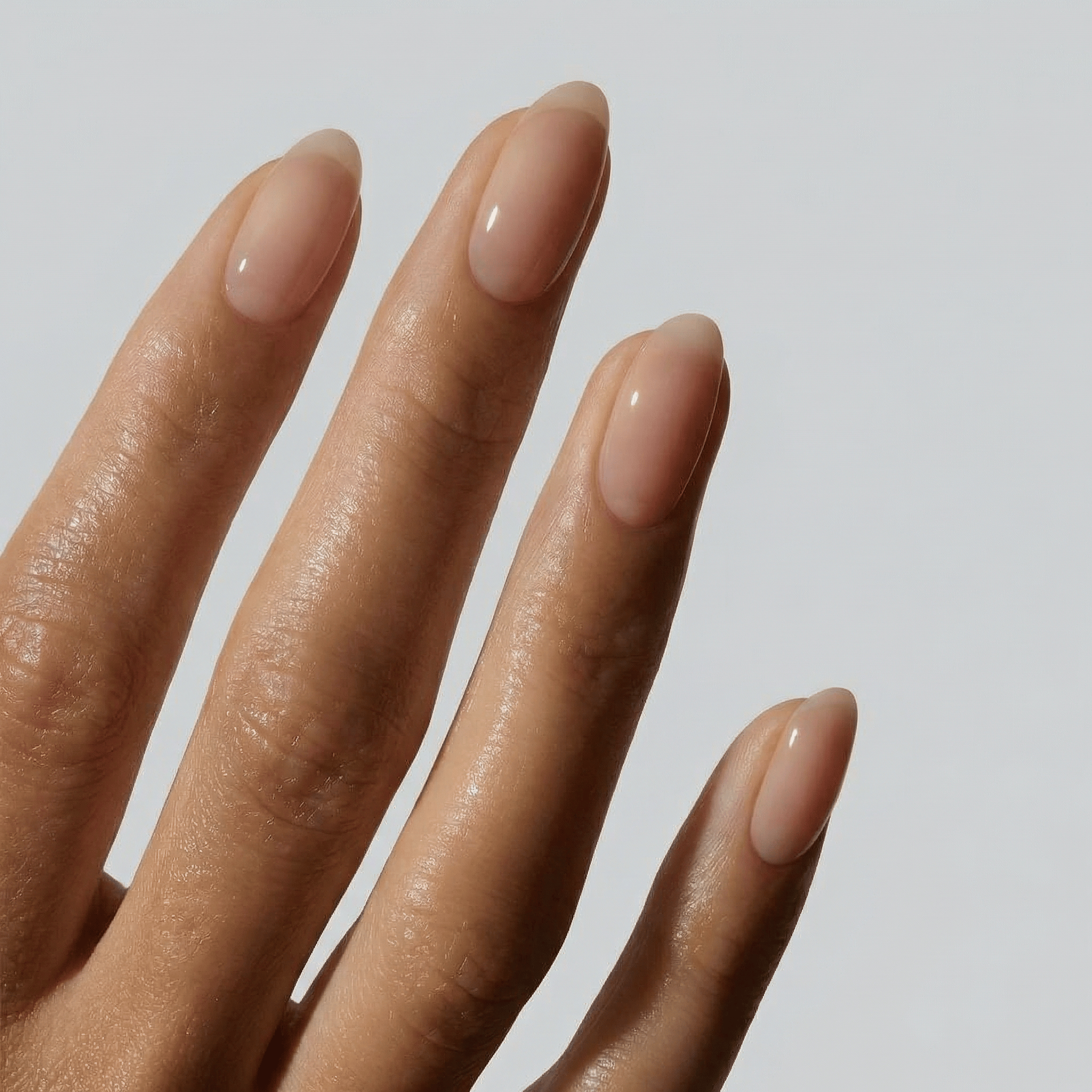 A close-up of a hand with neatly manicured, almond-shaped nails painted in a glossy, sheer nude pink gel polish against a light gray background.